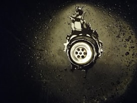 A metal sink drain surrounded by water droplets on a dark surface. The area around the drain has a reflective appearance with scattered droplets creating a glistening effect.