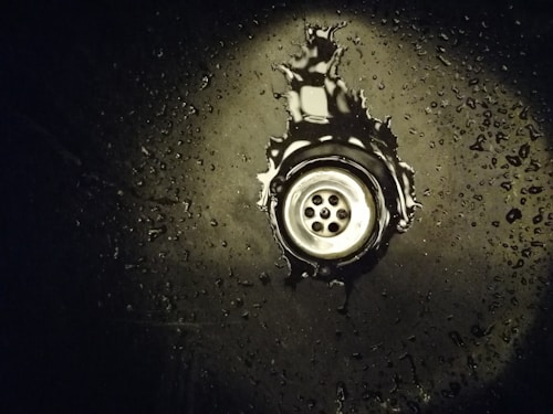 A metal sink drain surrounded by water droplets on a dark surface. The area around the drain has a reflective appearance with scattered droplets creating a glistening effect.