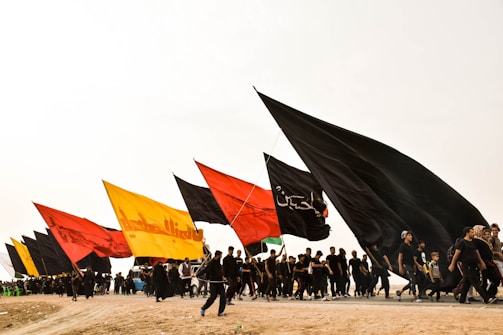 A group of people is walking together on a dirt path, holding large flags of various colors, including black, red, yellow, and green. The sky is overcast, and there are a number of people dressed in black, suggesting a somber or significant event.