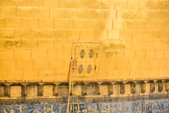 A wall covered in large, golden tiles with an intricately patterned small door in the center. Below, there is a decorative strip featuring geometric and possibly textual designs in blue and gold.
