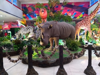 A vibrant display in a public space features large, realistic models of various animals including a rhinoceros, zebra, giraffe, and a roaring lion on a rock. The scene is set amidst lush green plants and colorful background art, creating a vivid and dynamic atmosphere.