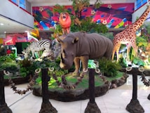 A vibrant display in a public space features large, realistic models of various animals including a rhinoceros, zebra, giraffe, and a roaring lion on a rock. The scene is set amidst lush green plants and colorful background art, creating a vivid and dynamic atmosphere.