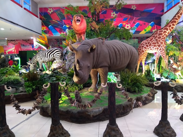 A vibrant display in a public space features large, realistic models of various animals including a rhinoceros, zebra, giraffe, and a roaring lion on a rock. The scene is set amidst lush green plants and colorful background art, creating a vivid and dynamic atmosphere.