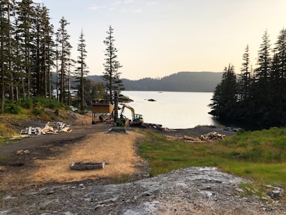 A serene lakeside scene with a dirt pathway leading to a small wooden structure. There is construction equipment nearby, surrounded by tall trees and a calm body of water in the background. The sky is slightly overcast, casting a soft light over the landscape.
