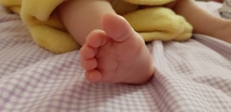 A small baby foot is tucked under a soft yellow blanket. The foot is resting on a light purple and white checkered fabric, likely a bed sheet. The image conveys a sense of warmth and comfort, highlighting the delicate nature of an infant.