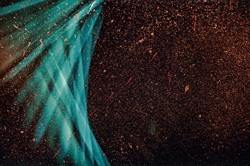 A dark background with scattered particles and light streaks intersecting. The particles appear to be floating or suspended, creating a sense of depth and movement. The light streaks are primarily blue-green and create a contrasting dynamic against the dark backdrop.