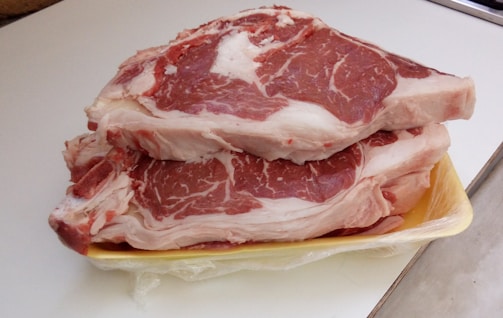 Two raw steaks with marbled fat are stacked on a yellow plastic tray. The steaks exhibit a mix of red meat interspersed with white fat, sitting on a countertop.