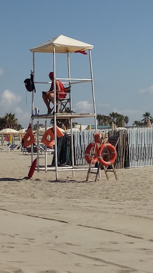 A lifeguard sits atop a tall white wooden lifeguard chair on a sandy beach. The lifeguard is wearing a red uniform and a red cap, observing the beach. Several orange lifebuoys are attached to the structure. In the background, there is a line of sun umbrellas and palm trees under a clear blue sky.