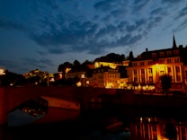 A picturesque evening scene of a quaint town with historic architecture. The buildings are illuminated with warm, golden lights that contrast against the deep blue sky and the water reflecting the scene. A bridge in the foreground serves as a focal point, adding to the charm and tranquility of the setting.