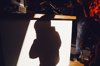 A shadow of a small child cast on a light-colored wall is prominently featured. The shadow suggests the child is standing with one hand near the face or in a contemplative pose. Surrounding the shadow, partially illuminated objects and a person's legs are visible, with a dimly lit environment in the background.