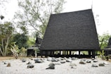 A traditional black Thai-style house with a steeply pitched roof made from dark wood shingles. Surrounding the structure are trees and bushes, and the foreground features a gravel area dotted with various smooth stones.