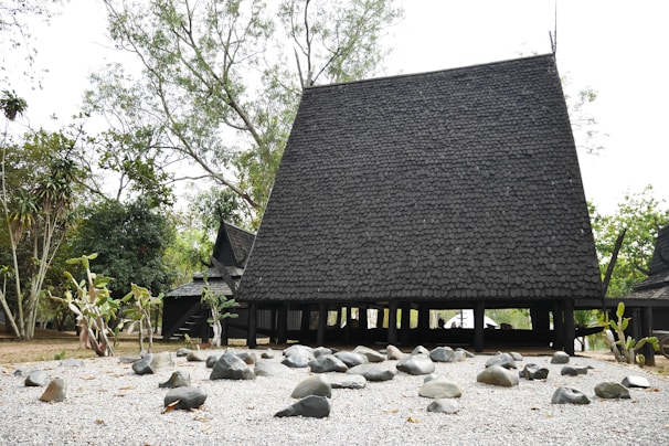 A traditional black Thai-style house with a steeply pitched roof made from dark wood shingles. Surrounding the structure are trees and bushes, and the foreground features a gravel area dotted with various smooth stones.