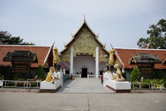 A traditional Thai temple with a richly decorated gable and intricate golden carvings is flanked by two large golden statues, likely mythical creatures, at the entrance. The temple features red-tiled roofs and lush greenery in the surroundings, with signs in Thai script.