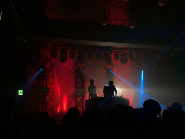 A dimly lit nightclub scene with two silhouetted DJs standing behind a console, surrounded by vibrant red and blue stage lights. The background features a series of spotlights and a light fog filling the space, creating a lively and energetic atmosphere.