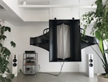 A modern living space featuring a large, abstract black sound system hanging from the ceiling. On the floor, there are two white standing speakers on either side, and a metal shelving unit housing electronic equipment. Potted plants with lush green leaves add a touch of nature to the minimalistic decor.