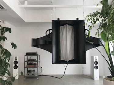A modern living space featuring a large, abstract black sound system hanging from the ceiling. On the floor, there are two white standing speakers on either side, and a metal shelving unit housing electronic equipment. Potted plants with lush green leaves add a touch of nature to the minimalistic decor.