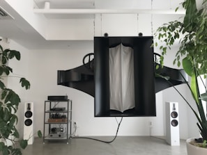 A modern living space featuring a large, abstract black sound system hanging from the ceiling. On the floor, there are two white standing speakers on either side, and a metal shelving unit housing electronic equipment. Potted plants with lush green leaves add a touch of nature to the minimalistic decor.
