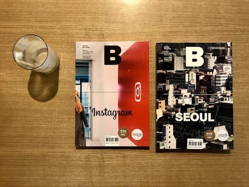 Two magazines with bold 'B' on their covers placed on a wooden table next to a nearly empty glass with some milk residue. The left magazine features 'Instagram' with a red and white cover, while the right one features 'Seoul' with an urban skyline photograph.