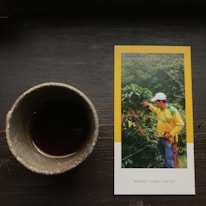 A ceramic cup filled with a dark liquid is placed on a wooden surface next to a photograph. The photograph features a person wearing a yellow and green shirt and a white cap, pointing towards coffee plants in a lush, green setting. 'Market Lane Coffee' is printed on the lower part of the photograph.