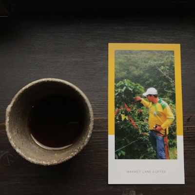 A ceramic cup filled with a dark liquid is placed on a wooden surface next to a photograph. The photograph features a person wearing a yellow and green shirt and a white cap, pointing towards coffee plants in a lush, green setting. 'Market Lane Coffee' is printed on the lower part of the photograph.