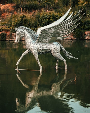 A metallic sculpture of a Pegasus with intricate details stands in a body of water, creating a reflection. Lush greenery surrounds the scene, adding a natural backdrop.