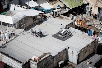 A rooftop with various objects including a reclining chair, a person seated in the chair, exercise equipment, and satellite dishes. The surrounding area includes makeshift roofing and various household items such as air conditioning units and a coiled hose.
