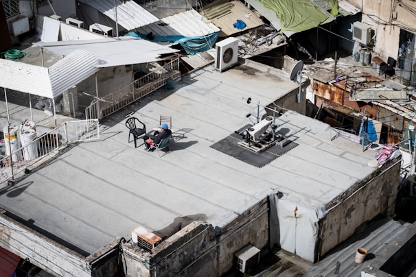 A rooftop with various objects including a reclining chair, a person seated in the chair, exercise equipment, and satellite dishes. The surrounding area includes makeshift roofing and various household items such as air conditioning units and a coiled hose.