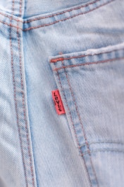 Close-up of a light blue denim fabric with visible red stitching and a small red tag sewn into the seam.