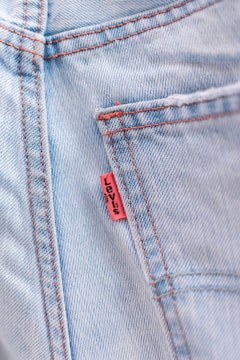 Close-up of a light blue denim fabric with visible red stitching and a small red tag sewn into the seam.