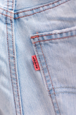 Close-up of a light blue denim fabric with visible red stitching and a small red tag sewn into the seam.