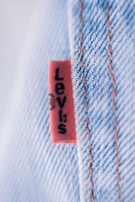 A close-up view of a denim fabric with prominent stitching details. A small red tag with black text is visible, partially showing a brand name. The fabric texture and stitching are clearly defined, illustrating the quality and craftsmanship of the material.