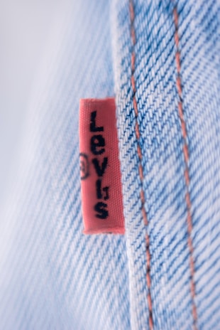 A close-up view of a denim fabric with prominent stitching details. A small red tag with black text is visible, partially showing a brand name. The fabric texture and stitching are clearly defined, illustrating the quality and craftsmanship of the material.
