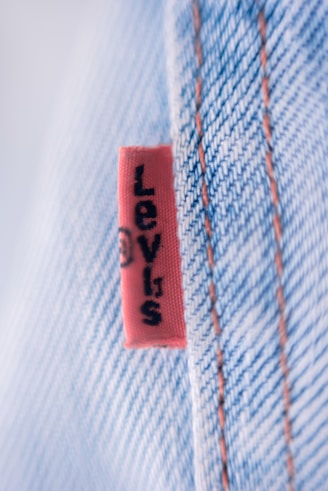 A close-up view of a denim fabric with prominent stitching details. A small red tag with black text is visible, partially showing a brand name. The fabric texture and stitching are clearly defined, illustrating the quality and craftsmanship of the material.