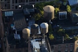 An aerial view of a rooftop in an urban setting featuring multiple cylindrical water towers. These structures are surrounded by greenery and various rooftop installations, including vents and utility boxes. The scene is bustling with urban life, as plants thrive around the installations, creating a mix of industrial and natural elements.