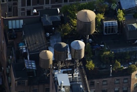 An aerial view of a rooftop in an urban setting featuring multiple cylindrical water towers. These structures are surrounded by greenery and various rooftop installations, including vents and utility boxes. The scene is bustling with urban life, as plants thrive around the installations, creating a mix of industrial and natural elements.