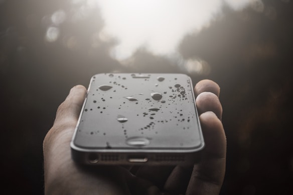 A hand holding a smartphone covered in water droplets against a blurred natural background. The focus is on the glossy surface of the phone where the water droplets are clearly visible, creating a reflective effect.