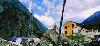 A mountainous landscape scene features a tall communication tower surrounded by lush green hills. In the foreground, solar panels are installed on a rocky surface. Two people stand on the rocks, gazing toward a mustard-colored building with dark windows. The sky is partly cloudy, casting a serene light on the scenery.