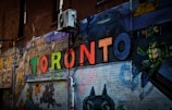 A vibrant street art piece painted on a brick wall featuring the word 'TORONTO' in colorful, bold letters. Surrounding the word are various cartoon-style characters, adding a playful and artistic atmosphere. An air conditioning unit is mounted on the brick wall, highlighting the urban setting.