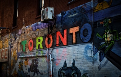 A vibrant street art piece painted on a brick wall featuring the word 'TORONTO' in colorful, bold letters. Surrounding the word are various cartoon-style characters, adding a playful and artistic atmosphere. An air conditioning unit is mounted on the brick wall, highlighting the urban setting.