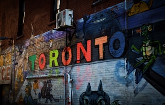 A vibrant street art piece painted on a brick wall featuring the word 'TORONTO' in colorful, bold letters. Surrounding the word are various cartoon-style characters, adding a playful and artistic atmosphere. An air conditioning unit is mounted on the brick wall, highlighting the urban setting.