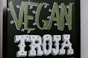 The image features a blackboard-style sign with bold lettering. The word 'VEGAN' is written in a light green color with small leaf accents around it, and the word 'TROJA' is below in bold white letters with a decorative font.