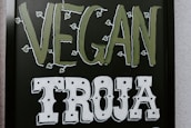The image features a blackboard-style sign with bold lettering. The word 'VEGAN' is written in a light green color with small leaf accents around it, and the word 'TROJA' is below in bold white letters with a decorative font.