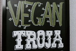 The image features a blackboard-style sign with bold lettering. The word 'VEGAN' is written in a light green color with small leaf accents around it, and the word 'TROJA' is below in bold white letters with a decorative font.