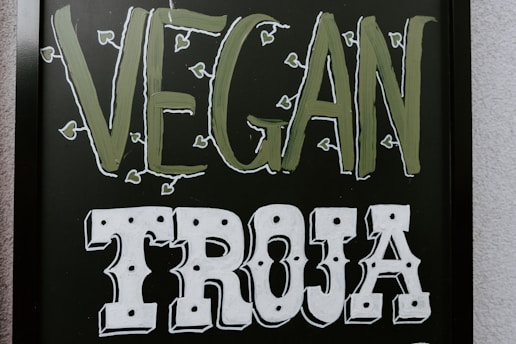 The image features a blackboard-style sign with bold lettering. The word 'VEGAN' is written in a light green color with small leaf accents around it, and the word 'TROJA' is below in bold white letters with a decorative font.