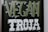 The image features a blackboard-style sign with bold lettering. The word 'VEGAN' is written in a light green color with small leaf accents around it, and the word 'TROJA' is below in bold white letters with a decorative font.