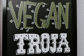 The image features a blackboard-style sign with bold lettering. The word 'VEGAN' is written in a light green color with small leaf accents around it, and the word 'TROJA' is below in bold white letters with a decorative font.