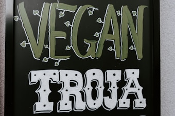 The image features a blackboard-style sign with bold lettering. The word 'VEGAN' is written in a light green color with small leaf accents around it, and the word 'TROJA' is below in bold white letters with a decorative font.