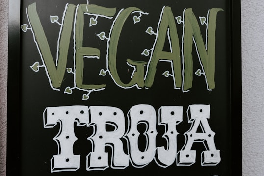 The image features a blackboard-style sign with bold lettering. The word 'VEGAN' is written in a light green color with small leaf accents around it, and the word 'TROJA' is below in bold white letters with a decorative font.