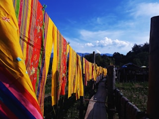 Brightly colored textiles with intricate patterns are hung on a line against a backdrop of clear blue skies and distant mountains. The textiles feature vibrant yellows, reds, and greens with floral and striped designs. A wooden pathway is visible below the textiles, and trees fill the surrounding landscape.