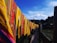 Brightly colored textiles with intricate patterns are hung on a line against a backdrop of clear blue skies and distant mountains. The textiles feature vibrant yellows, reds, and greens with floral and striped designs. A wooden pathway is visible below the textiles, and trees fill the surrounding landscape.